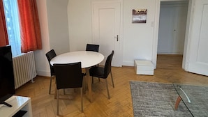 Et spiseområde med et hvidt bord, fire stole, en radiator og en dør med et lille trin til et andet rum.