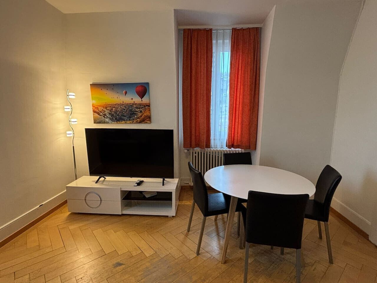Una habitación con una televisión de pantalla plana sobre un soporte blanco, una mesa de comedor con cuatro sillas, un cuadro de globos aerostáticos y una ventana con cortinas rojas.
