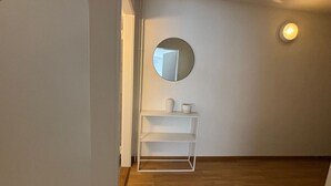 A hallway with a white shelf unit, a round mirror, and a light fixture on the wall.