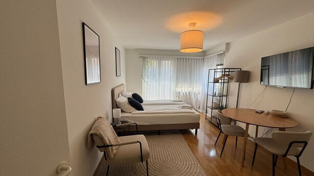 A modern hotel room with a bed, a desk, and a chair. There is a window with curtains and a lamp on the ceiling.