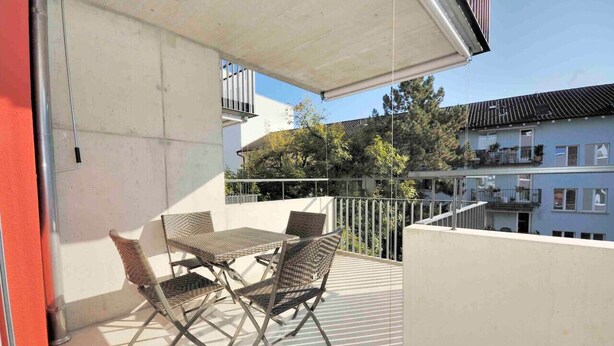 Un balcón con una mesa y sillas, barandilla de metal y vistas a edificios y árboles al fondo.