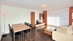 Una sala moderna con una mesa de comedor de madera, sillas negras, un sofá blanco, una mesa de centro de vidrio y un televisor de pantalla plana sobre un soporte blanco.