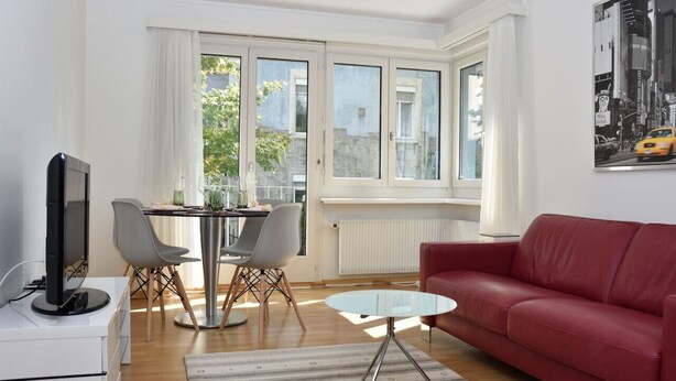 Een moderne woonkamer met een rode bank, een glazen salontafel, een eettafel met stoelen, en een televisie op een witte kast.