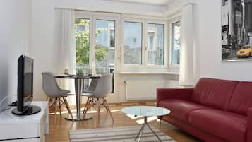 Ein modernes Wohnzimmer mit einem roten Sofa, einem Glastisch, einem Esstisch mit Stühlen und einem Fernseher auf einer weißen Kommode.