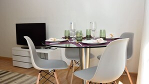 Een eetgedeelte met een glazen tafel, vier grijze stoelen, een flatscreen televisie en een witte kast.