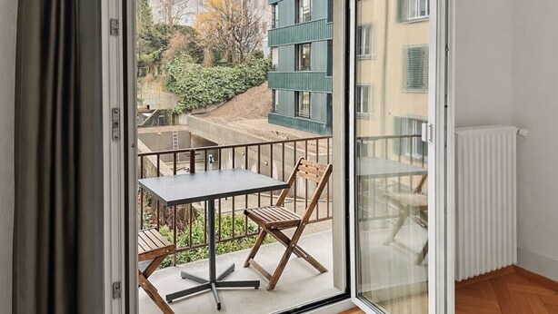A balcony with a table and chair, a view of buildings, and a window. - 2BR near lake (Stardust 11) (Zürich)