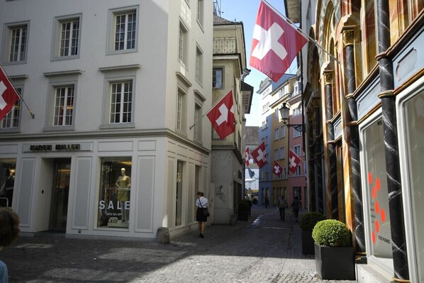 Une rue pavée bordée d’immeubles, dont certains arborent des drapeaux suisses. On peut voir une devanture de magasin avec une pancarte indiquant « SOLDES ».