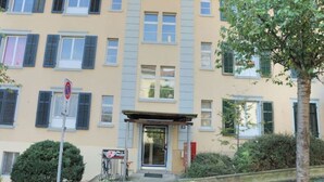 Un immeuble résidentiel à plusieurs étages, doté d’une façade beige, de volets bleus et d’un petit hall d’entrée surmonté d’un auvent.