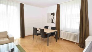 Un salon moderne avec un parquet en bois disposé en chevrons, une table basse en verre, un canapé beige, une table à manger avec quatre chaises et un radiateur.