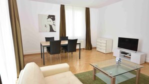 Un salon moderne avec une table basse en verre, un canapé beige, un coin repas avec une table et des chaises, un meuble blanc et un téléviseur à écran plat.