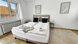 Una cama bien hecha con almohadas y mantas, una mesita de noche con lámpara y dos cuadros enmarcados en la pared.