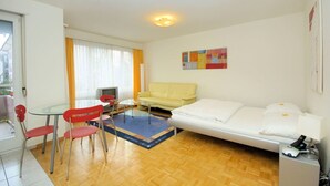A compact hotel room with a bed, a sofa, a dining table with chairs, a TV, and a small desk.