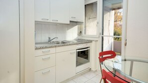A compact kitchen with white cabinets, a granite countertop, a stainless steel sink, and a built-in oven. There is a red chair and a glass table near a sliding glass door leading to a balcony.