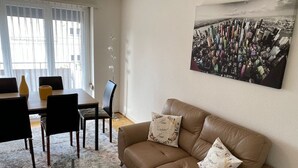 A living room with a brown leather sofa, two matching cushions, a glass coffee table, and a dining area with a black table and chairs.