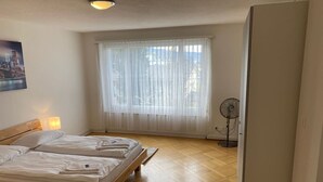 A bedroom with two beds, a window with sheer curtains, a standing fan, and a wooden floor.