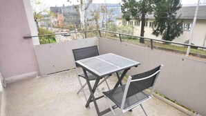 A balcony with a metal table and two chairs, a concrete floor, and a view of buildings and trees.