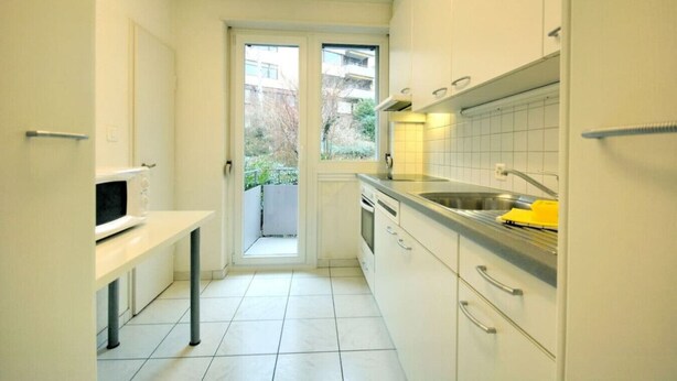 A compact kitchen with white cabinets, a microwave, a sink, and a window leading to a balcony.