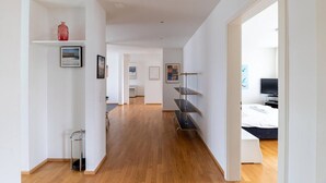 A spacious, well-lit hallway with wooden flooring, leading to a bedroom with a bed and a TV.