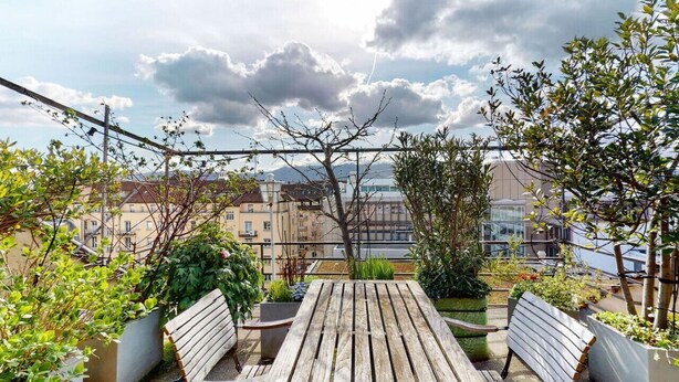 A balcony with wooden furniture, potted plants, and a view of buildings and a body of water.