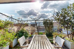 Een balkon met houten meubels, plantenbakken en uitzicht op gebouwen en een waterpartij.