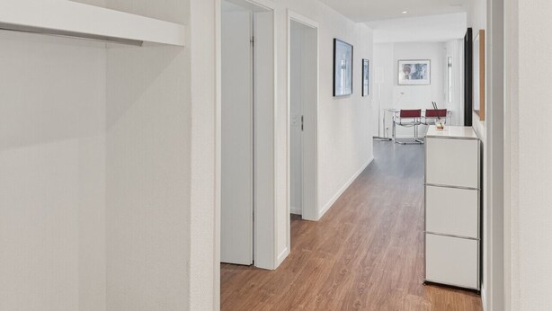 Ein Flur mit weißen Wänden, Holzboden und einem weißen Wandschrank. Eine Tür führt in einen anderen Raum, in der Ferne steht ein kleiner Tisch mit Stühlen.