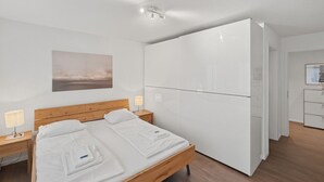 Ein modernes Hotelzimmer mit einem hölzernen Bett, weißer Bettwäsche, einem weißen Kleiderschrank und einem kleinen Nachttisch mit einer Lampe.