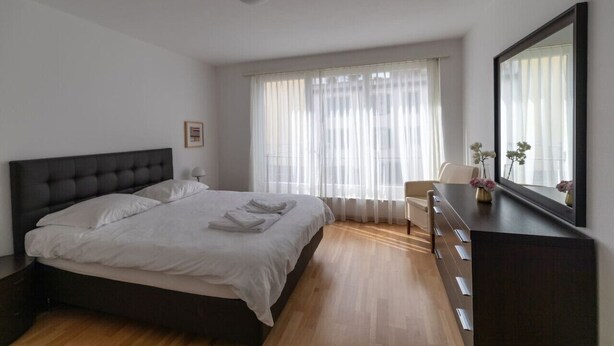Ein Schlafzimmer mit einem großen Bett, einem Schminktisch, einem Sessel und einem Fenster mit durchsichtigen Vorhängen.