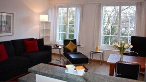 Ein modernes Wohnzimmer mit einer schwarzen Couch, einem Couchtisch, einem Beistelltisch mit Lampe und Blick durch große Fenster.