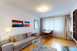 A modern living room with a sofa, coffee table, and dining table. There are two paintings on the wall, a lamp, and a large window with curtains. - Executive 1BR flat, city center (Hammer 1) (Zürich)