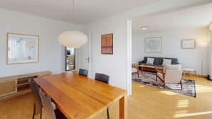 A spacious living area with a wooden dining table, a sofa, and a coffee table. There are framed pictures on the walls and a large window providing natural light.