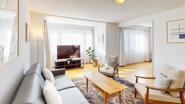 A modern living room with a sofa, armchair, and coffee table. There is a TV and a plant in the room.