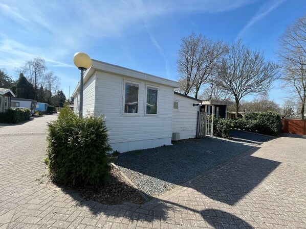 Une petite maison mobile d’un seul étage, dotée d’une antenne parabolique, d’une allée en gravier et d’un petit arbuste au premier plan.