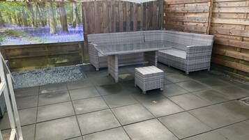 Un espace extérieur aménagé en terrasse, avec un canapé d'angle en rotin, une table à plateau en verre et un petit pouf.