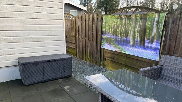 Une terrasse dotée d’une table à plateau de verre et de meubles en osier, d’une clôture en bois et d’un grand écran diffusant une scène de forêt.