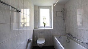 A modern bathroom with a toilet, bathtub, and shower. There is a window with a plant on the windowsill. - Bright 1 BR flat (Parkview 1) (Zürich)