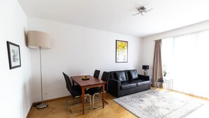 A living room with a wooden floor, a black leather sofa, a dining table with chairs, a lamp, and a large window with curtains. - Bright 1 BR flat (Parkview 1) (Zürich)