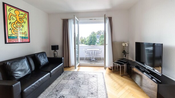 A modern living room with a black leather sofa, a flat-screen TV, and a patterned rug. - Bright 1 BR flat (Parkview 1) (Zürich)