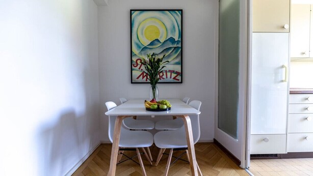 A dining area with a white table, four chairs, a vase with flowers, and a painting on the wall.