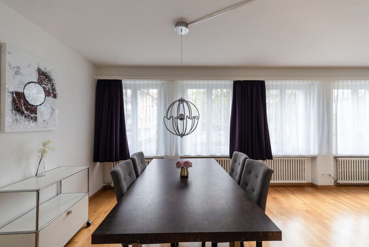 A modern dining room with a long table, dark curtains, a decorative light fixture, and a radiator.