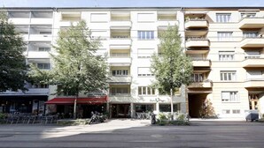 A multi-story building with balconies, a storefront with outdoor seating, and trees in front. - Amazing studio (37 sqm) city center/Enge (Wille 4) (Zürich)