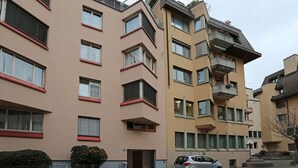 Un edificio residencial de varios pisos con balcones, un coche estacionado adelante y un pequeño portón de acceso.