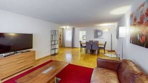 Una sala de estar moderna con televisor de pantalla plana, muebles de madera, un tapete rojo, un sofá de cuero y un comedor con mesa y sillas.