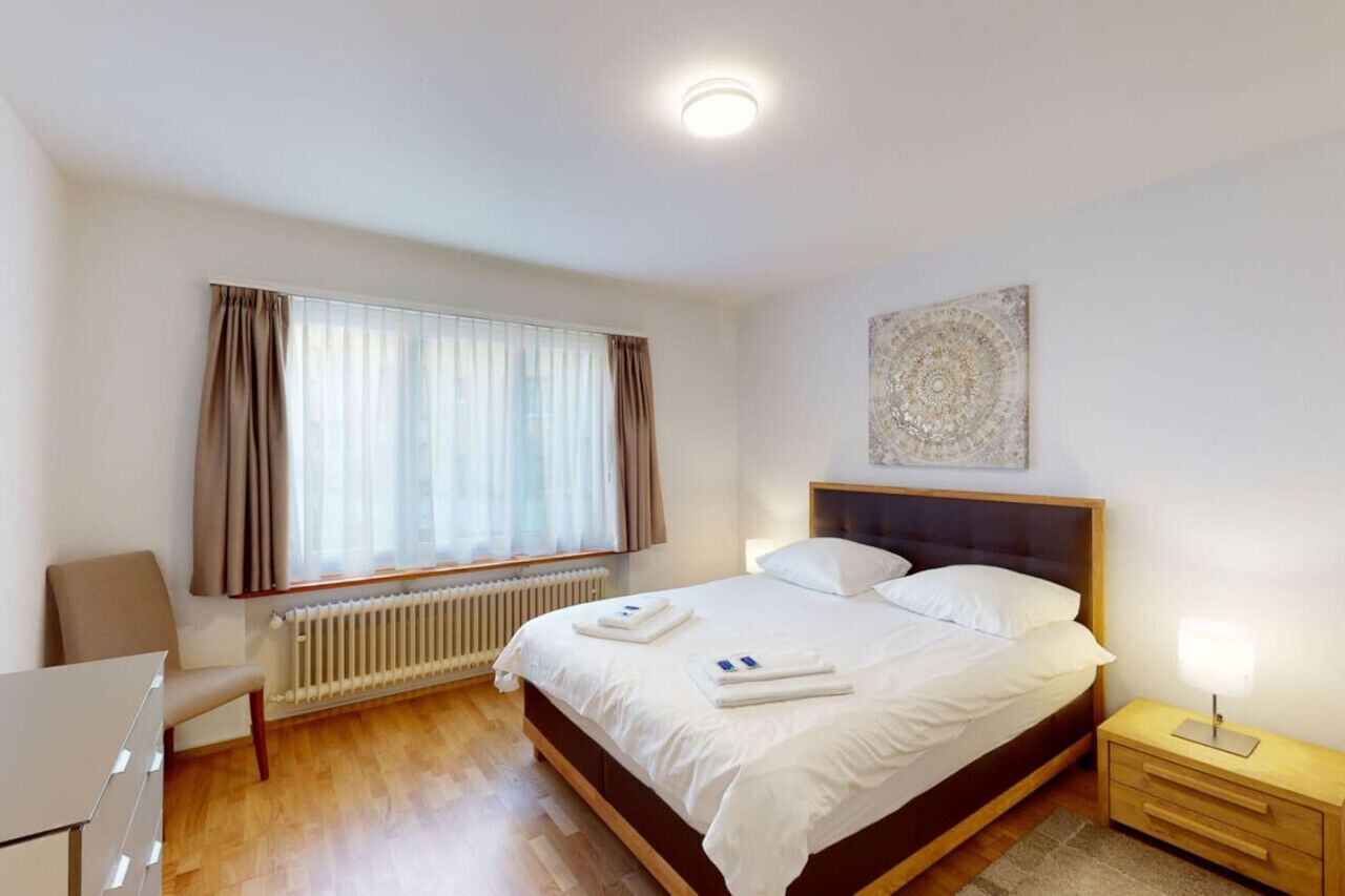 Une chambre à coucher comprenant un lit, une chaise, une table de chevet, un radiateur et une fenêtre avec des rideaux.