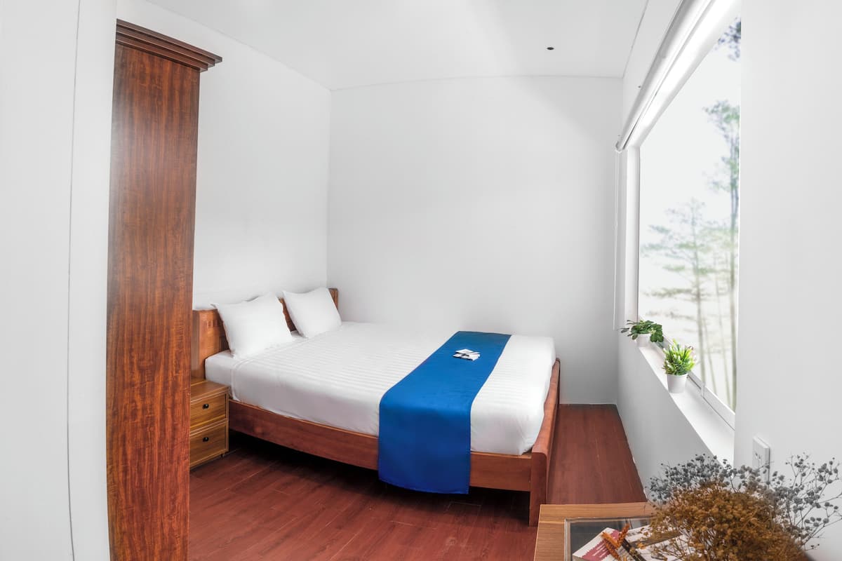 Un dormitorio con cama de madera, ropa de cama blanca, detalles en azul, una mesita de noche, un armario de madera, una ventana con plantas y vistas a árboles.
