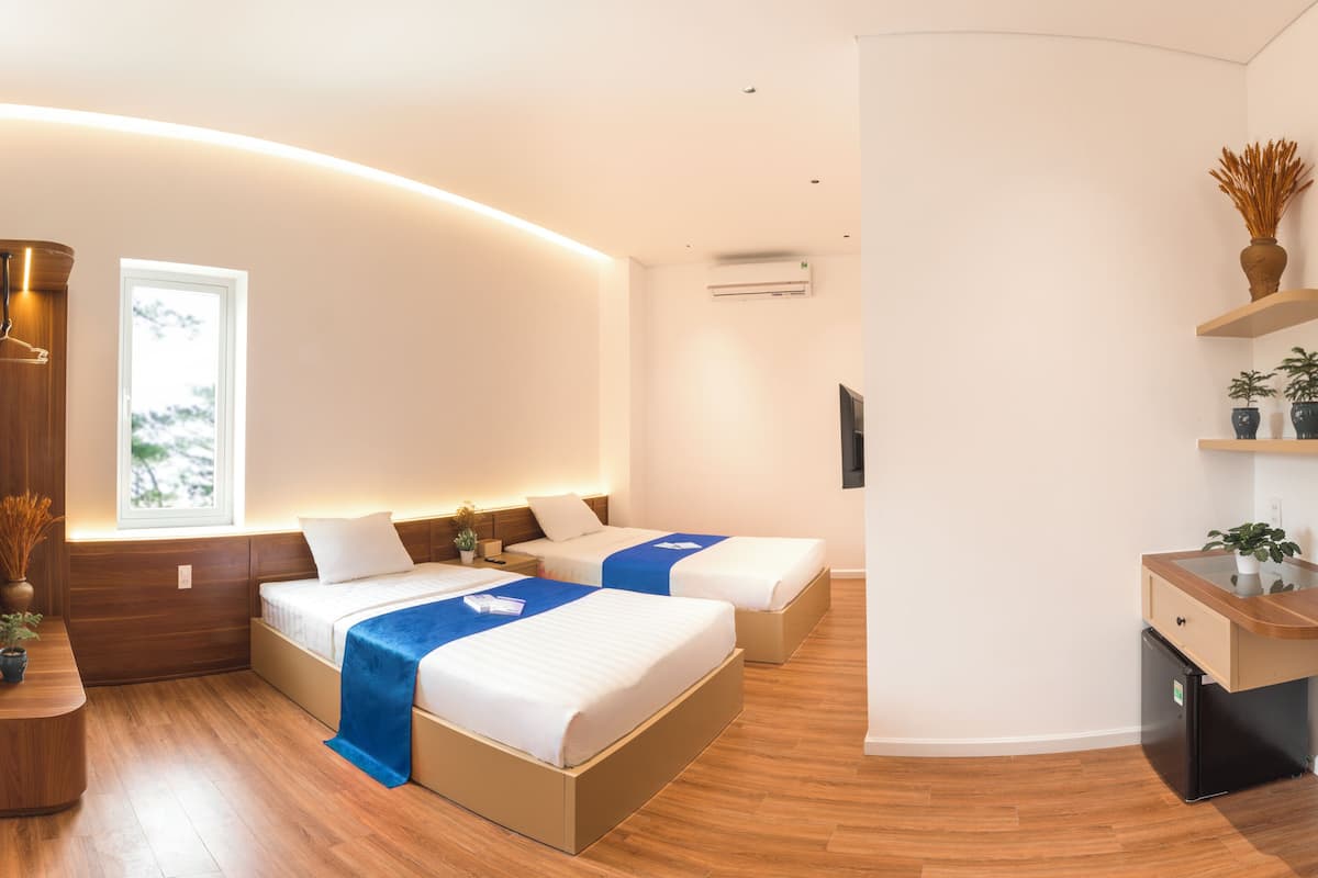 Una habitación de hotel moderna con dos camas, un mostrador de recepción de madera, una pequeña cocina y un gran ventanal que proporciona luz natural.