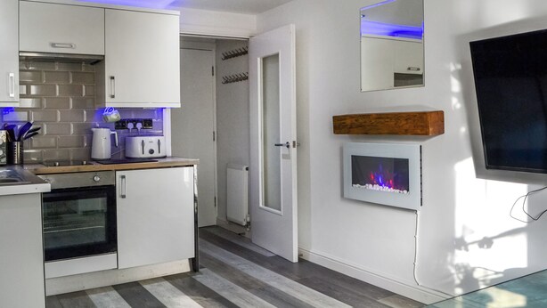 A modern kitchen with white cabinets, a built-in oven, and a coffee maker. There is a fireplace with a stone surround and a mirror above it.