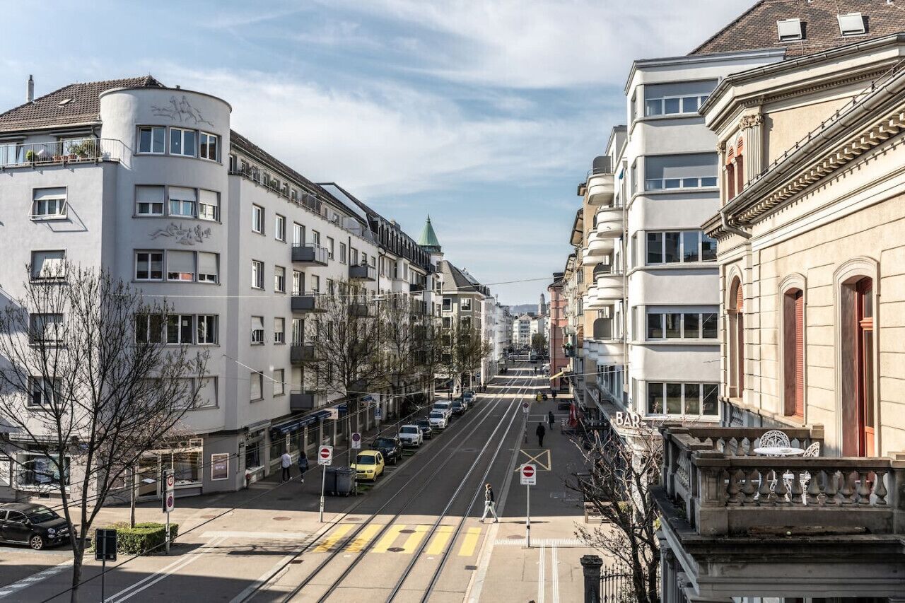 Straßenansicht mit Gebäuden, einer Straßenbahnhaltestelle und einer Fußgängerquerung.