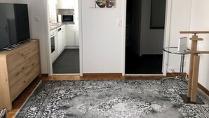 Une pièce comprenant une commode en bois, un téléviseur à écran plat, un tapis à motifs, une œuvre d’art accrochée au mur et une porte donnant sur la cuisine.