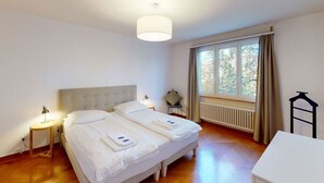 A double bed with white linens, a bedside table with a lamp, a window with curtains, and a radiator.