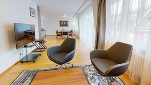 Ein modernes Wohnzimmer mit Flachbildfernseher, zwei Sesseln, einem Couchtisch und einem Essbereich mit Tisch und Stühlen.
